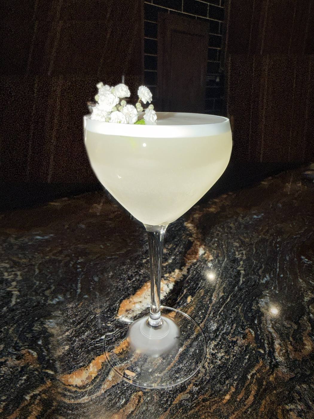 A white foamy cocktail in a coupe glass garnished with baby&rsquo;s breath on a granite bar