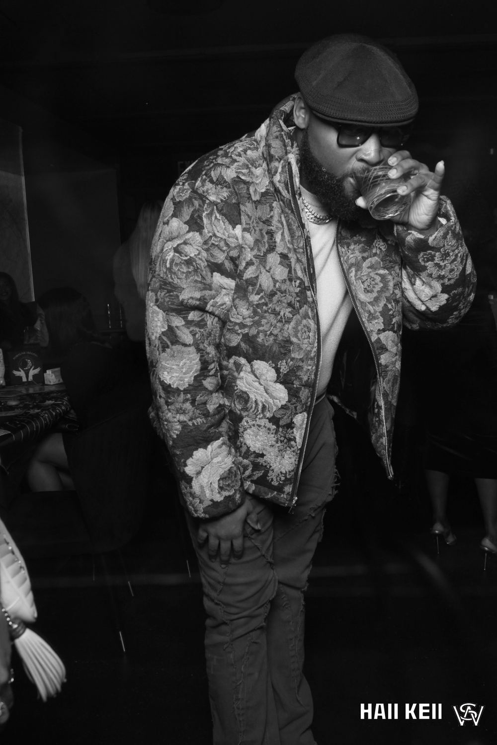 Black and white portrait of Self in a floral jacket and cap, sipping from a glass
