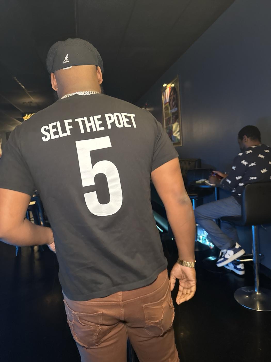 Self from behind in a black Self The Poet number 5 shirt and Kangol cap